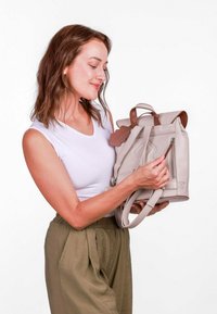 Světle šedý kožený batoh s hnědými akcenty, horním klopním víkem, nastavitelnými popruhy a zipovou kapsou na boku. Kompaktní a strukturovaný design.