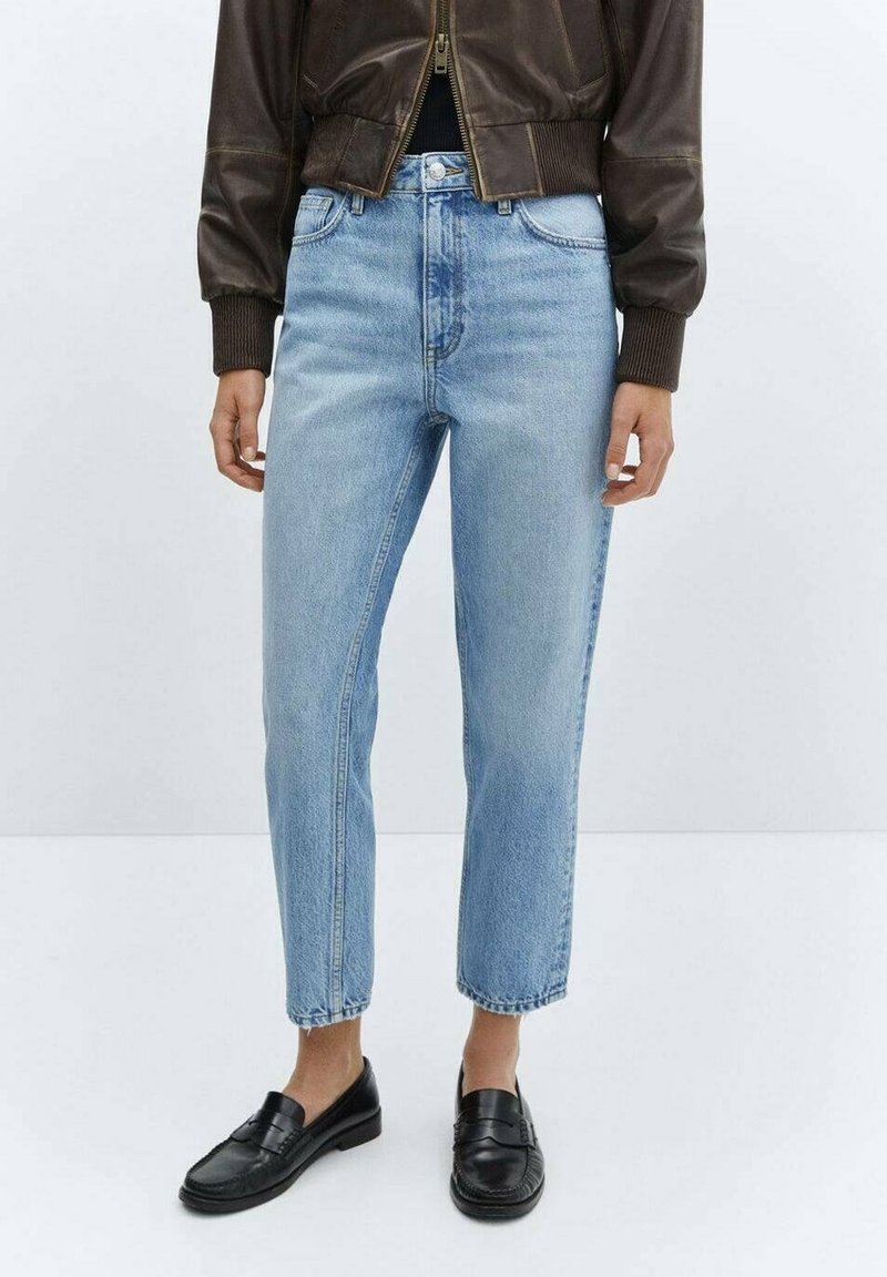 Personne portant un jean taille haute bleu clair, une veste en cuir marron courte et des mocassins noirs, debout sur un sol blanc devant un fond uni.