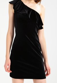 Černé šaty na jedno rameno s volánkovým akcentem. Vyrobeno z měkké sametové látky. Úzký střih a délka do kolen.