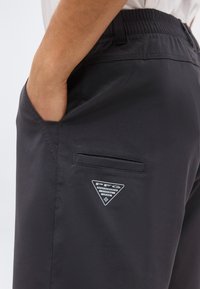 Άτομο που φορά σκούρα γκρι σορτς με το χέρι στην πλαϊνή τσέπη, εμφανίζοντας ένα μικρό τριγωνικό λογότυπο "PFG Performance Fishing Gear" στον μηρό.