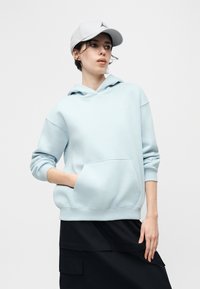 Νεαρό άτομο που φοράει γαλάζιο φούτερ με κουκούλα και λευκό καπέλο, στέκεται με το ένα χέρι στην τσέπη και κοιτάζει στο πλάι μπροστά σε μονόχρωμο φόντο.