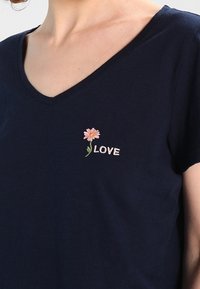 Námořnická modrá V-krční tričko s krátkými rukávy, zdobené růžovou květinovou výšivkou a slovem "LOVE" v bílé barvě, umístěným na levé části hrudi.
