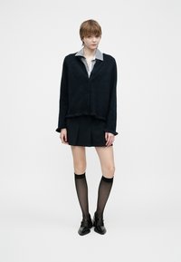 Mladý člověk stojí, oblečený v černém huňatém kardiganu přes košili s límečkem, černé plisované mini sukni, průhledných podkolenkách a černých botách.