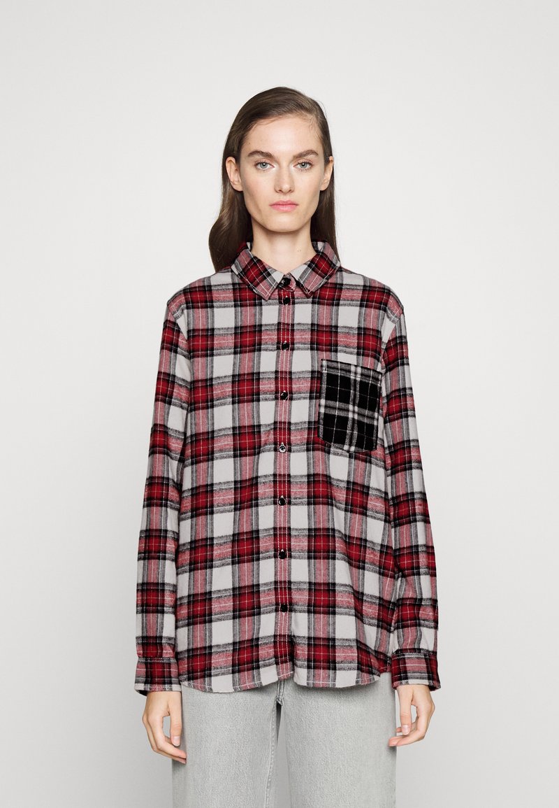 Kobieta ubrana w czerwoną i szaro-w kratkę koszulę zapinaną na guziki, z kontrastującą czarną kieszenią w kratkę na piersi, stojąca na tle białego tła.