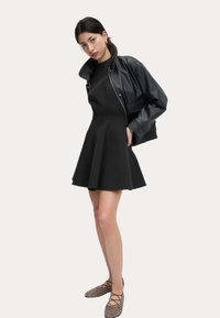 Schwarzes A-Linien-Kleid mit tailliertem Oberteil, strukturiertem Stoff und schwingendem Rock, kombiniert mit einer schwarzen Leder-Bomberjacke und Ballerinas im Leopardenmuster.