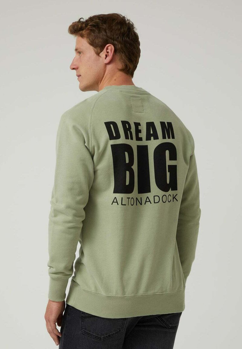 Hombre vistiendo un sudadera verde claro con el texto negro "DREAM BIG ALTONADOCK" en la parte de atrás, combinada con jeans oscuros, mirando de lado.