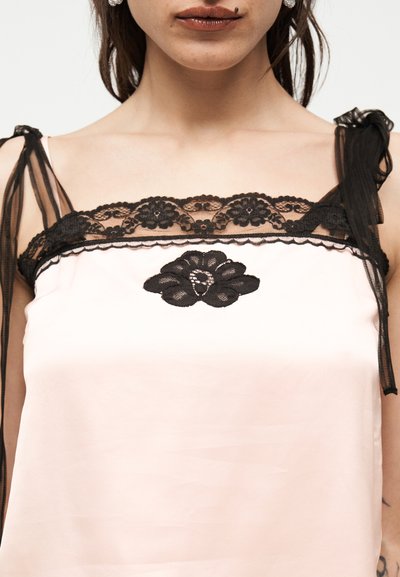 Femme portant un caraco rose pâle avec des bordures en dentelle noire et des bretelles à nœud, arborant un motif floral en dentelle noire sur la poitrine.