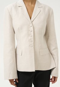 Béžový upravený blazer so štruktúrovaným strihom, s klapacím golierom, šiestimi okrúhlymi tlačidlami a bočnými vreckami, vyrobený z textúrovaného materiálu.