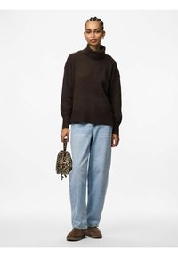 Maglione a collo alto marrone, vestibilità rilassata, orlo a coste. Jeans a gamba larga blu chiaro. Tiene in mano una piccola borsa a stampa leopardata. Scarpe slip-on marroni.
