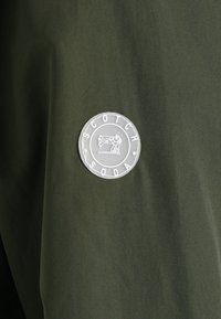 Olivengroene stof met een cirkelvormig logo waarop de tekst "SCOTCH SODA" en een naaimachineafbeelding in het wit staan. Soepele textuur, modern ontwerp.