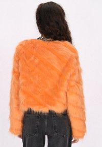 Orange fuskpälsjacka med kort längd och långa ärmar, med en mjuk, fluffig textur och en subtil randig mönstring. Kombinerad med svarta byxor.