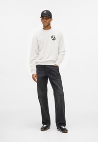 Fiatal férfi fehér logós pulóverben, fekete sapkában, fekete farmerben és fekete sportcipőben áll egy sima fehér háttér előtt.