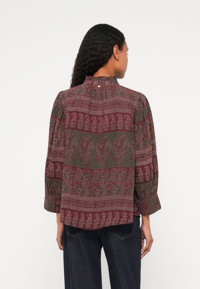 Sarkans un zaļš rakstains blūze ar augstu apkakli, pūkainām piedurknēm un pogu dekoru aizmugurē. Audumam ir ziedu un ģeometriski raksti.