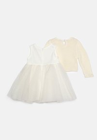 Ljusbeige klänning med figurnära överdel, tyllkjol och glittrande detaljer, i kombination med en kort, stickad krämfärgad kofta med öppning i ryggen.