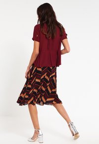 Blouse bordeaux à manches courtes et plissée, associée à une jupe plissée arborant des motifs géométriques en rouge, beige et marine. Talons blocs argent.
