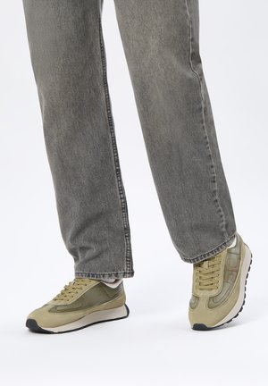 Beine in locker sitzenden grauen Jeans und olivgrünen Turnschuhen mit weißen Mittelsolen und schwarzen Gummiaußensohlen, stehend auf einer weißen Oberfläche.