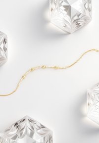 Collana d'oro con perle rotonde lisce e accenti di cristallo trasparente su una catena delicata, esposta su uno sfondo chiaro con oggetti di vetro.