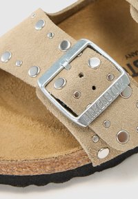 Sandale en daim beige avec des accents en clous argentés, une boucle métallique et un fond en liège avec une semelle noire texturée.