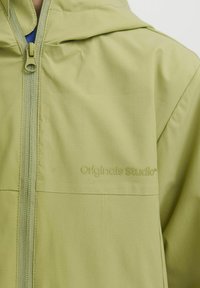 Ljust grön zip-up-jacka i texturerad tyg, med huva och tonalt varumärke "Originals Studio" på framsidan.