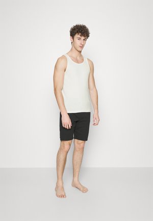 Weißes Tanktop aus weichem Stoff, kombiniert mit schwarzen Shorts. Das Tanktop hat einen runden Ausschnitt und breite Träger.