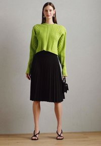 Pull cropped en maille côtelée vert vif, jupe midi plissée noire, sac noir et sandales à talons. Détails en maille texturée et en tissu fluide.