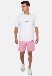 Maglietta bianca in cotone con logo nero, abbinata a pantaloni corti rosa chiaro e scarpe bianche con dettagli rossi. Abbigliamento estivo casual.