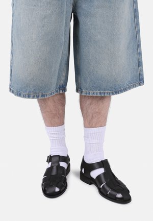 Jambes portant un short en denim bleu clair, des chaussettes blanches montantes et des sandales fermées en cuir tressé noir sur un fond uni.