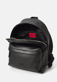 Zaino in pelle nera con tasca frontale con zip, scomparto principale parzialmente aperto che mostra l'etichetta rossa "HUGO" all'interno.