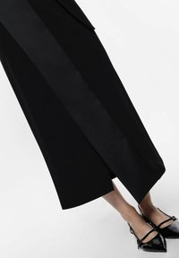 Abito nero su misura con una finitura satinata elegante, dotato di uno spacco laterale e di un design minimalista, abbinato a tacchi a punta in vernice nera.
