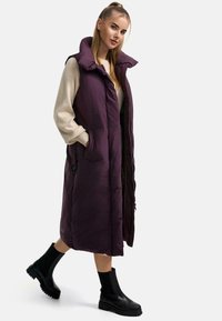 Gilet piumino lungo viola con colletto alto, zip frontale, tasche laterali e una texture lucida, abbinato a stivaletti neri alla caviglia.
