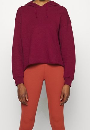 Osoba ubrana w bordową bluzę z kapturem i legginsy w kolorze spalanej pomarańczy, stojąca na jednolitym jasnym tle z rękami swobodnie opuszczonymi wzdłuż ciała.