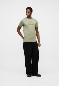 Homme portant un t-shirt vert clair "KARL LAGERFELD", un pantalon ample noir et des chaussures noires, debout avec une main derrière le dos.