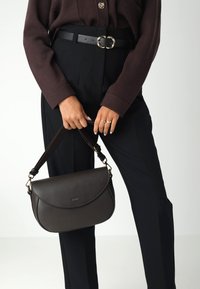 Persona che indossa un maglione marrone scuro e pantaloni neri, mentre tiene una borsa in pelle marrone scuro con dettagli in oro all'altezza della vita.