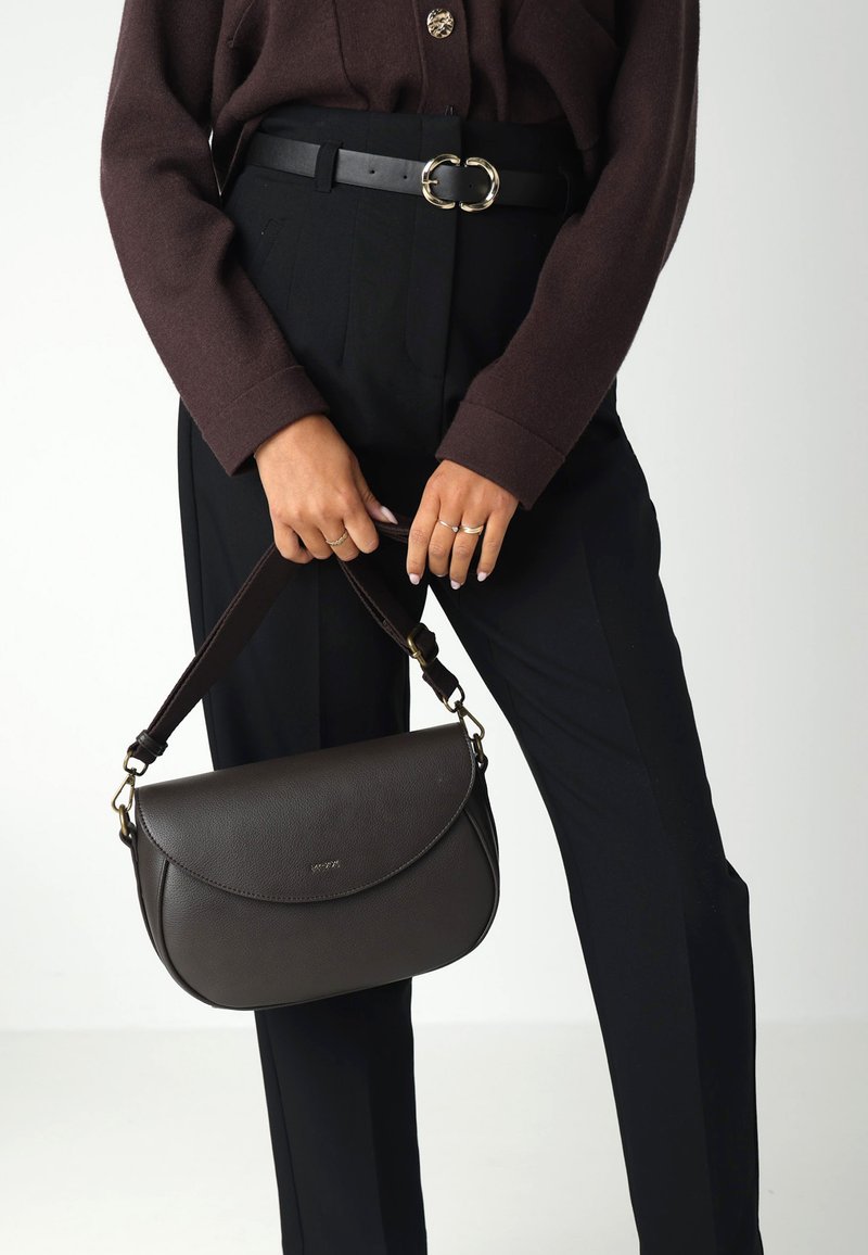 Persona che indossa un maglione marrone scuro e pantaloni neri, mentre tiene una borsa in pelle marrone scuro con dettagli in oro all'altezza della vita.