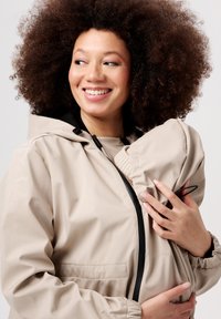 Beige vattentät jacka med huva, med elastiska ärmslut och dragkedja. Justeras för att bära ett spädbarn bekvämt. Slät textur.