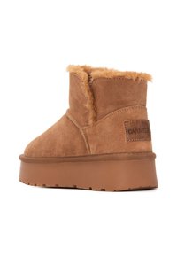 Botas de tobillo de ante marrón con forro de piel de peluche, suela de goma plana y una etiqueta de cuero con la marca en el lado. Punta redondeada, diseño sin costuras.