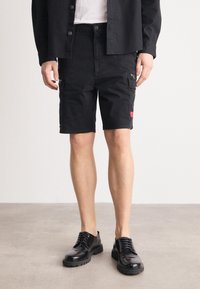 Man i svarta knapp-up shorts med sidofickor och svarta polerade snörskor, står på ett slätt golv.