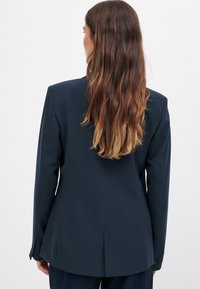 Ciemnoniebieska marynarka dopasowana, ze strukturalnym krojem, pojedynczym rozporkiem z tyłu i zapięciami na guzikach na mankietach. Wykonana z gładkiego materiału o subtelnej teksturze.