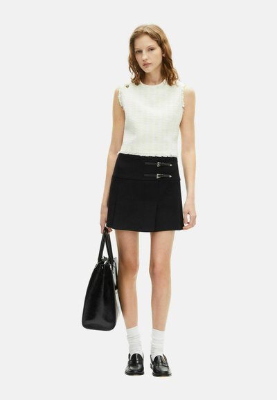 Mladá žena v bezrukávovom bielom topu, čiernej mini sukni s prackami, bielych ponožkách, čiernych mokasínach a drží čiernu koženú kabelku.