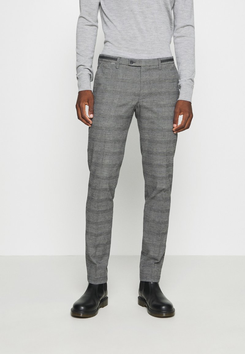 Homme portant un pantalon gris à carreaux, une chemise gris clair à manches longues et des bottes en cuir noires, debout sur un fond blanc uni.
