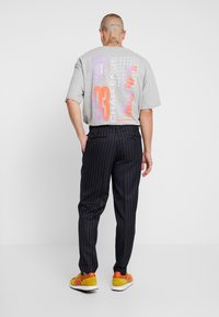 Grå grafisk t-shirt med orange och lila design, svarta randiga byxor och gula sneakers med röda detaljer. Enfärgad bakgrund.