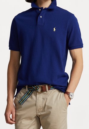 Mand iført en blå kortærmet polo, beige bukser med et stribet stofbælte og et armbåndsur, hænder afslappet ned langs siden.