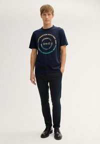 T-shirt en coton bleu marine avec un design graphique circulaire comportant du texte et des chiffres. Associé à un pantalon sombre et des chaussures à lacets noires.