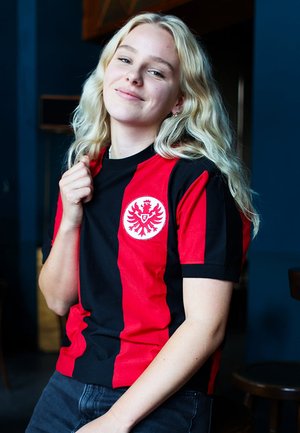Jonge persoon met lang blond haar die een rood-zwart sportshirt met een adelaarsembleem draagt, glimlacht en de kraag van het shirt vasthoudt.