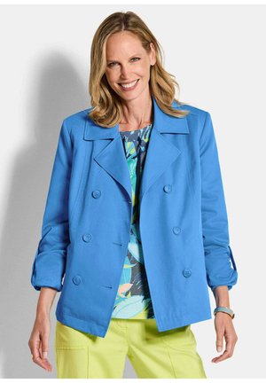 Glunderende vrouw die een felblauw double-breasted jasje, een blauw bedrukt topje en gele broek draagt, staand tegen een witte achtergrond.