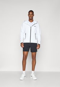 Weißer Reißverschlussjacke mit schwarzen Akzenten, Kapuze und Logo; kombiniert mit schwarzen Shorts und weißen Turnschuhen. Glatter Stoff und athletischer Schnitt.