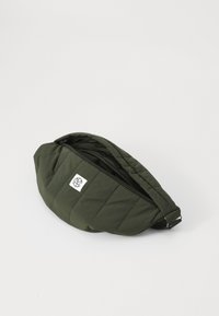 Gequilte olijfgroene heuptas met zwarte rits, witte logopatch en verstelbare strap. Compacte, ronde vorm, gladde textuur.