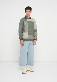 Giovane in maglione verde con pattern, pantaloni corti larghi di jeans azzurri chiari, calzini bianchi e sneakers beige, in piedi contro uno sfondo bianco.