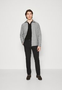 Chaqueta de cremallera gris claro con tela texturizada sobre una camiseta polo negra, combinada con pantalones negros y zapatos marrón oscuro.
