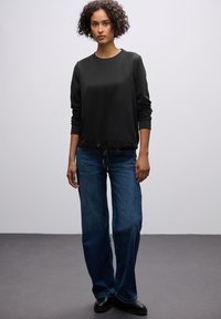 Sudadera negra con mangas acampanadas y dobladillo con cordón, combinada con jeans de pierna ancha azul y zapatos negros, sobre un fondo gris.